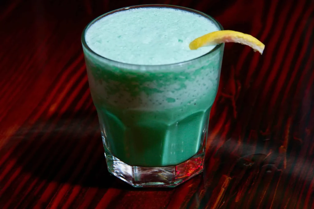 Frothy, layered green drink in a faceted glass with citrus garnish.