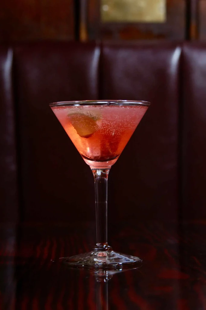 Reddish-pink effervescent cocktail in martini glass, citrus wedge, dark background.