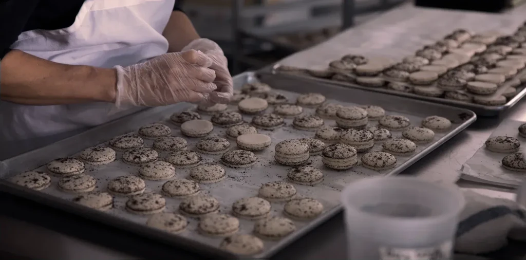 Gloved hands assemble dozens of light macarons on baking sheets.
