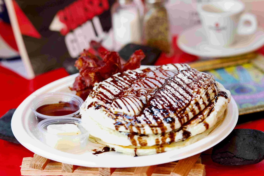 Tall stack of fluffy pancakes with chocolate sauce, cream, powdered sugar, bacon.