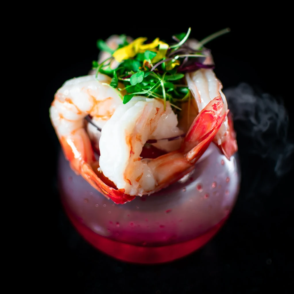 Peeled shrimp, microgreens, and red misty drink in a bulbous glass.