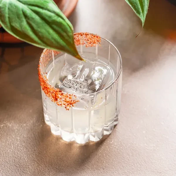 Close-up cocktail with embossed ice cube, spiced rim, and green leaf.