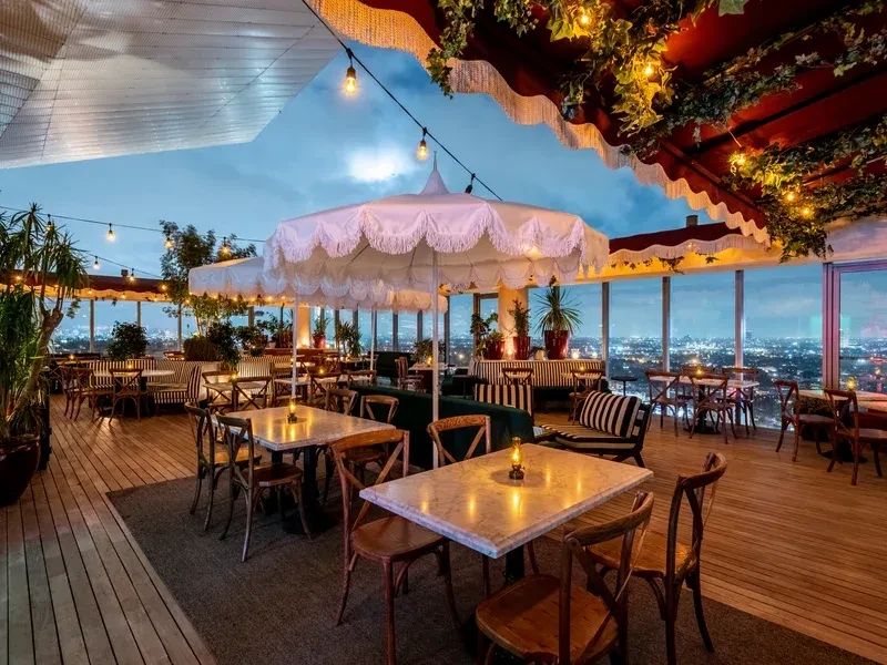 Empty rooftop dining area with tables, umbrella, string lights, and cityscape.