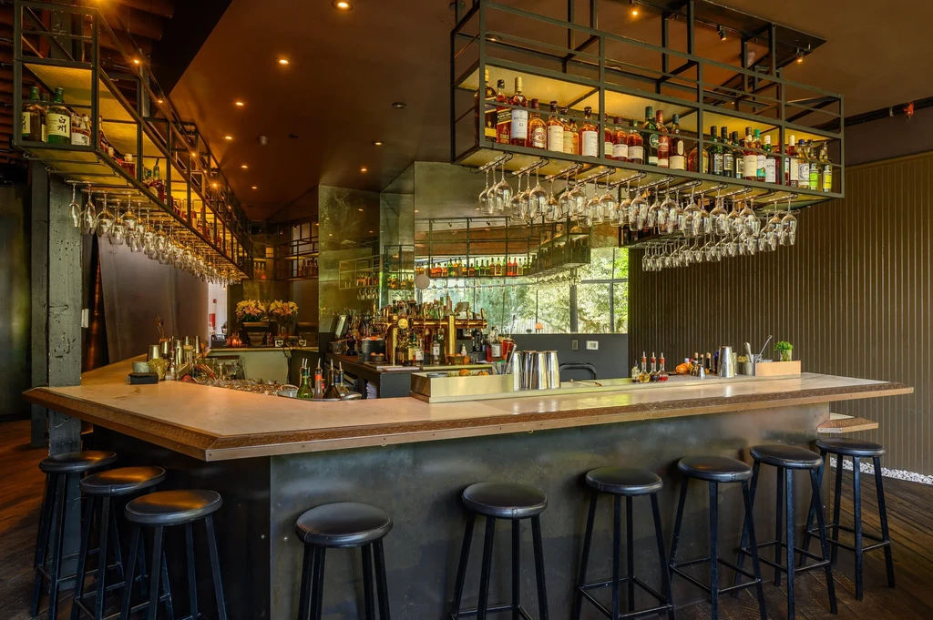 Empty, modern bar with L-shaped counter, many bottles, and hanging glasses.