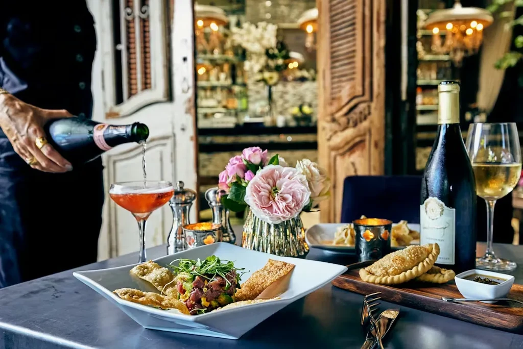 Hand pouring rosé at elegant restaurant table with tuna and empanadas.