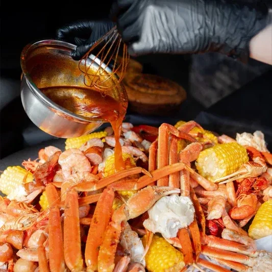 Gloved hand pours thick sauce over vibrant crab, shrimp, corn seafood boil.