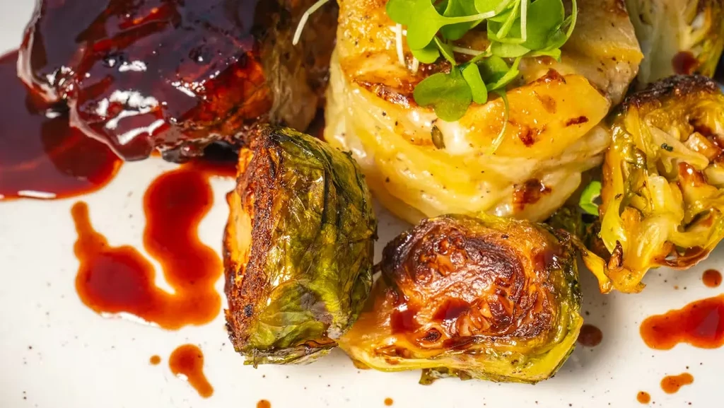 Plated dish with glazed meat, potato gratin, Brussels sprouts, and microgreens.