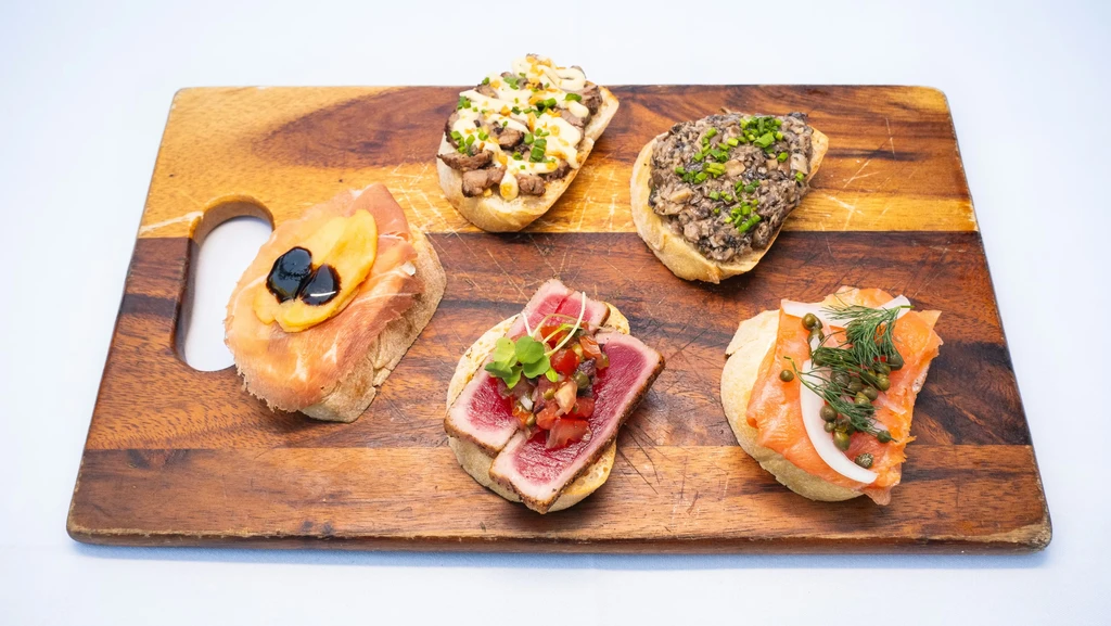 Five distinct bruschetta-style appetizers on a rustic wooden board.