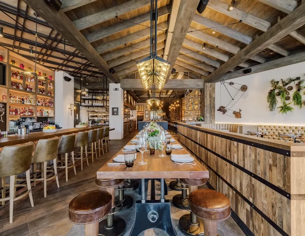 Modern restaurant interior with vaulted wooden ceiling, communal table, and bar.