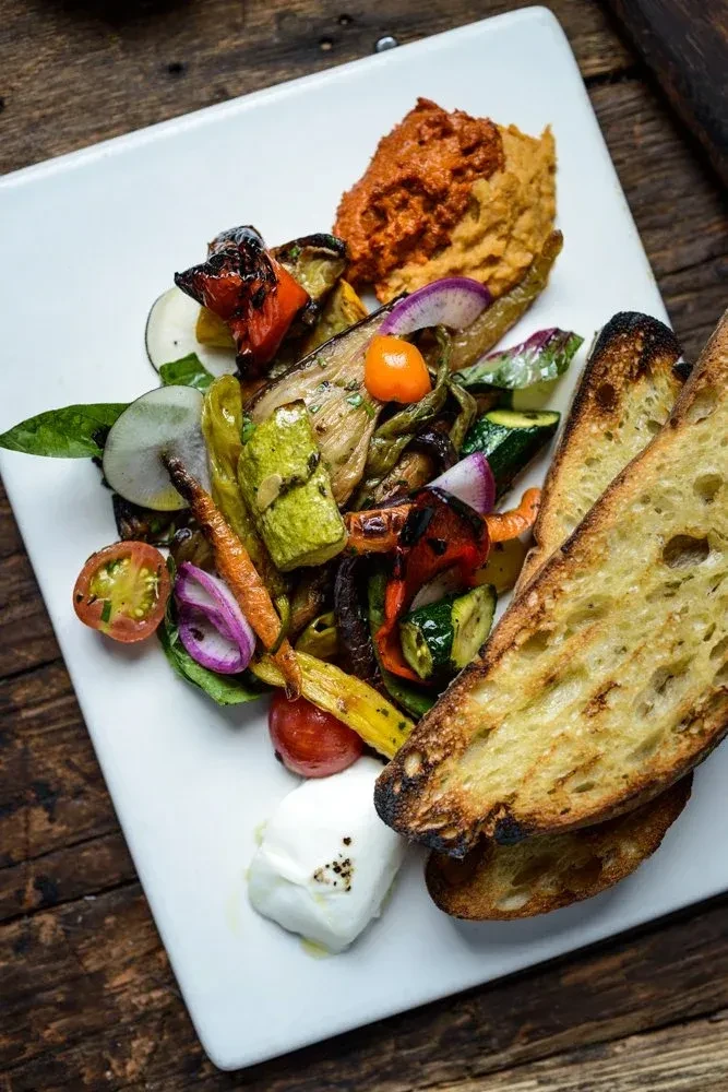 Plated roasted vegetables, hummus, mozzarella, and grilled bread.