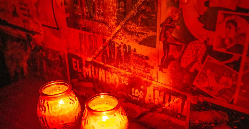 Two flickering candles illuminate a red-lit room with collage walls.
