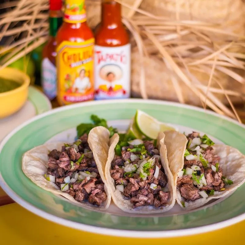 Three beef tacos with onion, cilantro, lime wedges, and hot sauces.