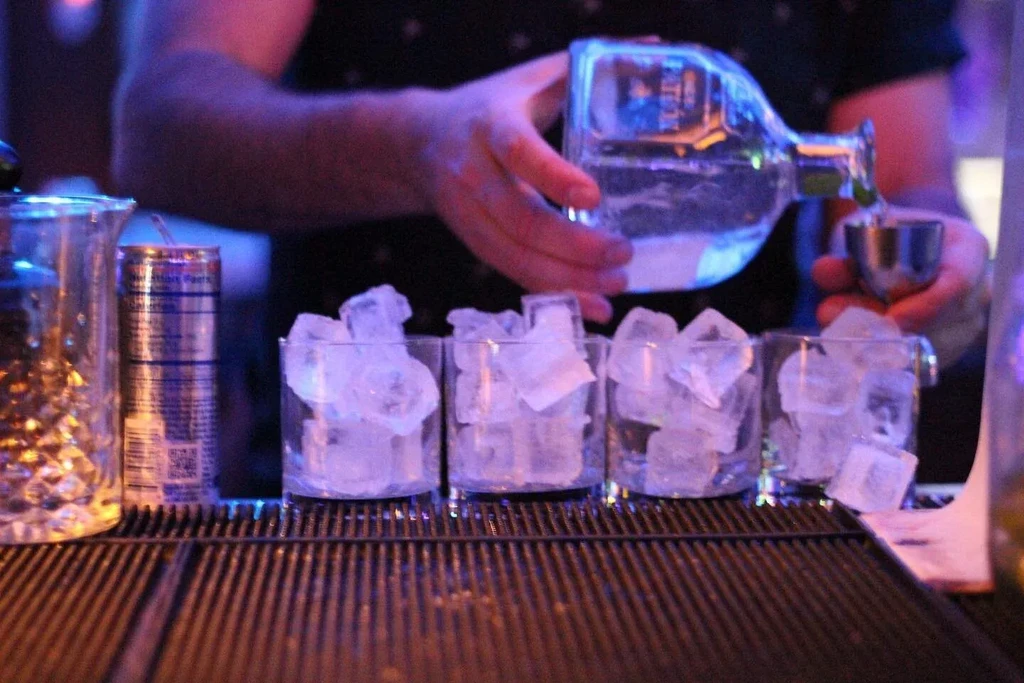 Bartender's hands pour liquor into jigger, ice-filled glasses, purple light.