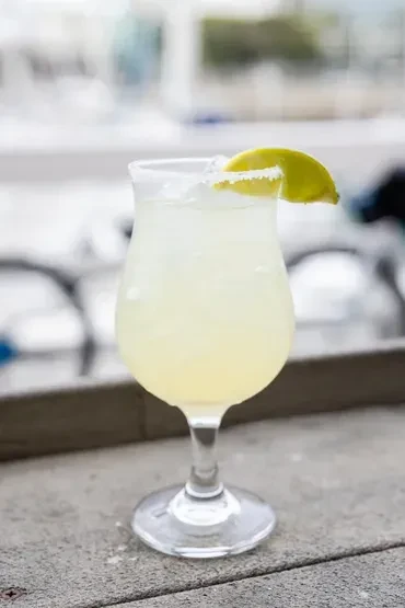 Close-up of a stemmed, salt-rimmed, pale yellow cocktail with lime.