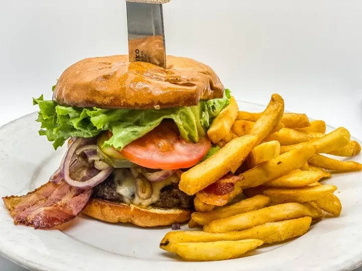 Close-up of a burger with melted cheese, bacon, knife, and fries.