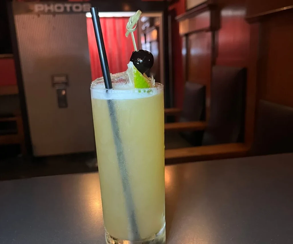 A tall garnished cocktail with foam, straw, near a photobooth.