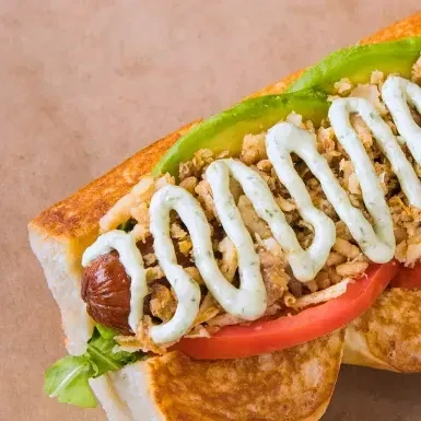 Gourmet hot dog in toasted bun with avocado, crispy onions, tomato, sauce.