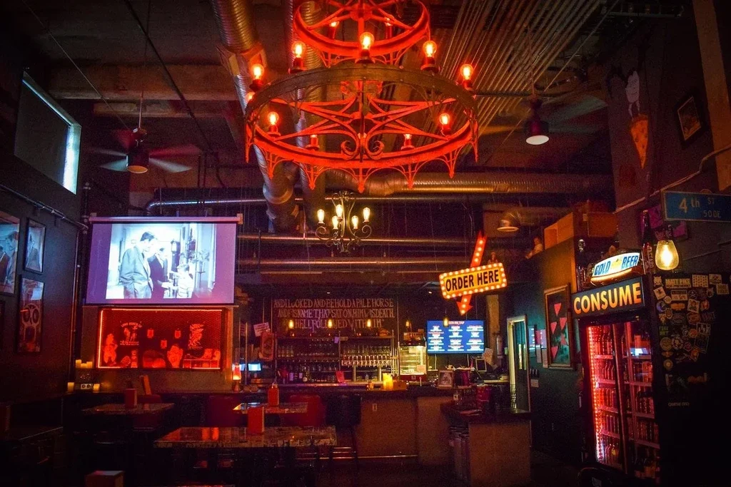 Dimly lit industrial-vintage bar interior with a red chandelier and movie.
