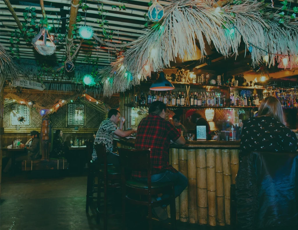 Interior of a dim, cozy tiki bar with bamboo counter and patrons.
