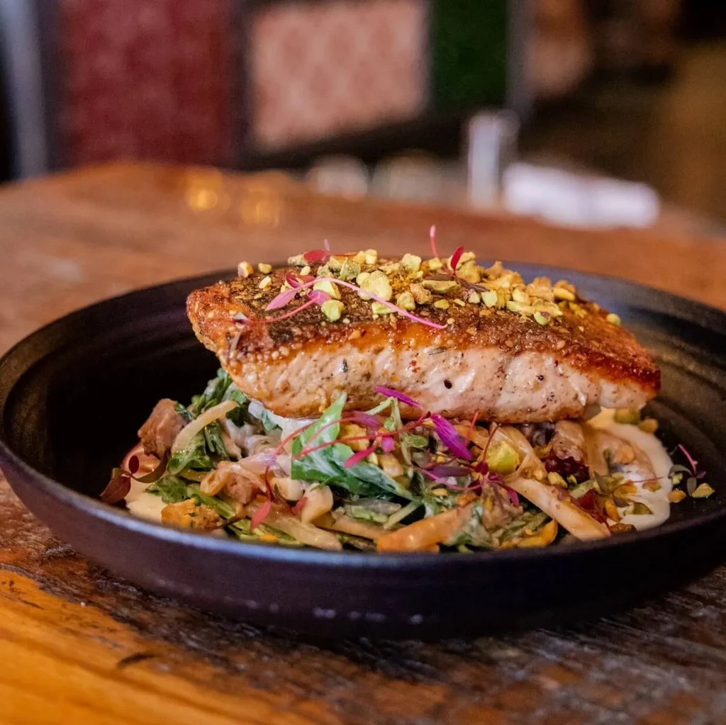 Seared salmon with pistachios, microgreens, greens, and sauce in a bowl.