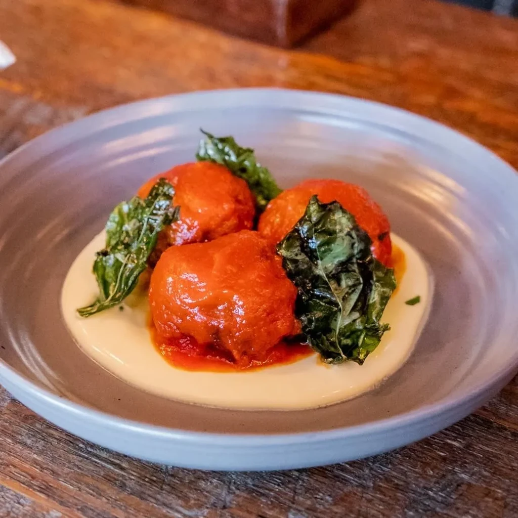 Four rounded items in reddish-orange sauce on white puree, green garnish.