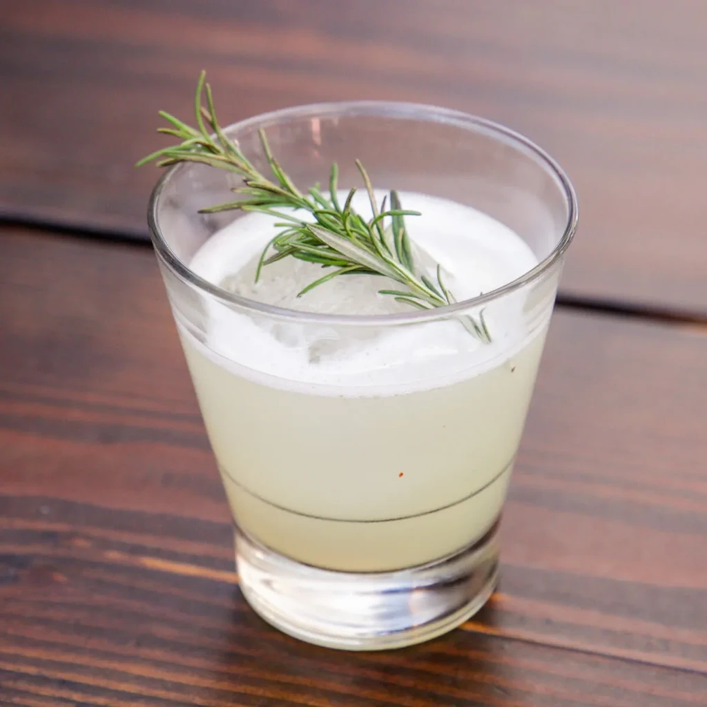 Light yellowish-green cocktail with foam, rosemary, and spherical ice.