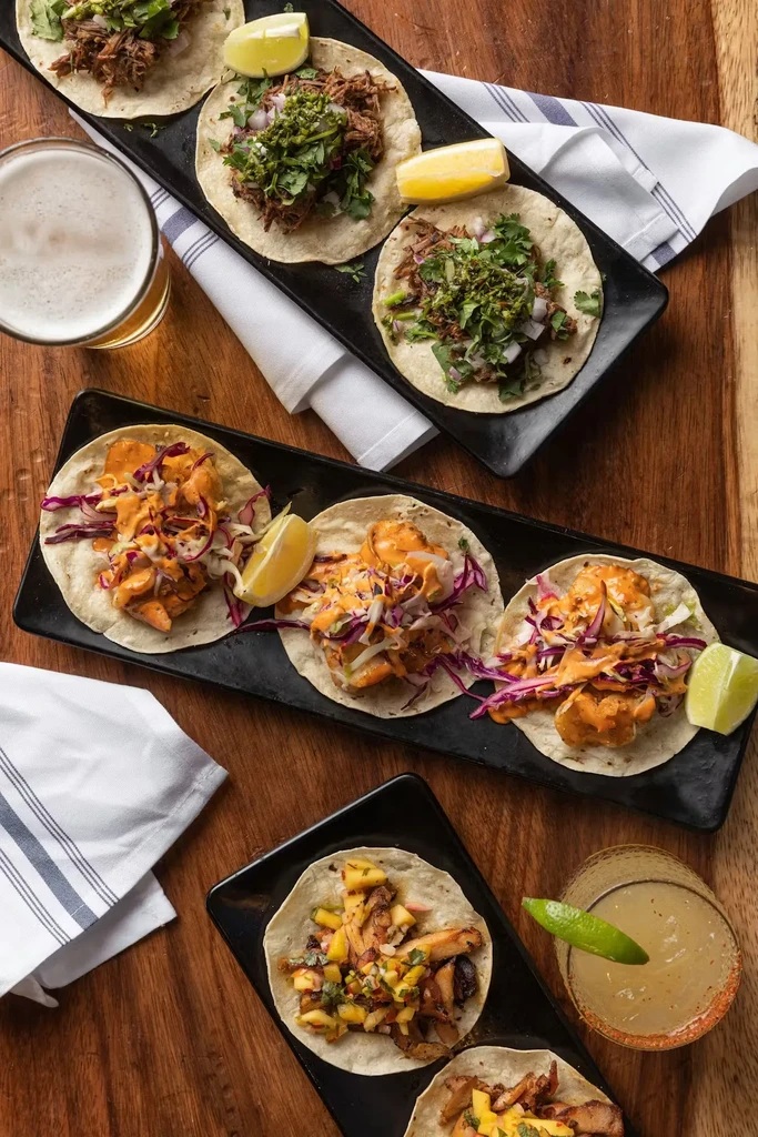 Overhead view: diverse tacos, beer, and cocktail on a rustic wooden table.