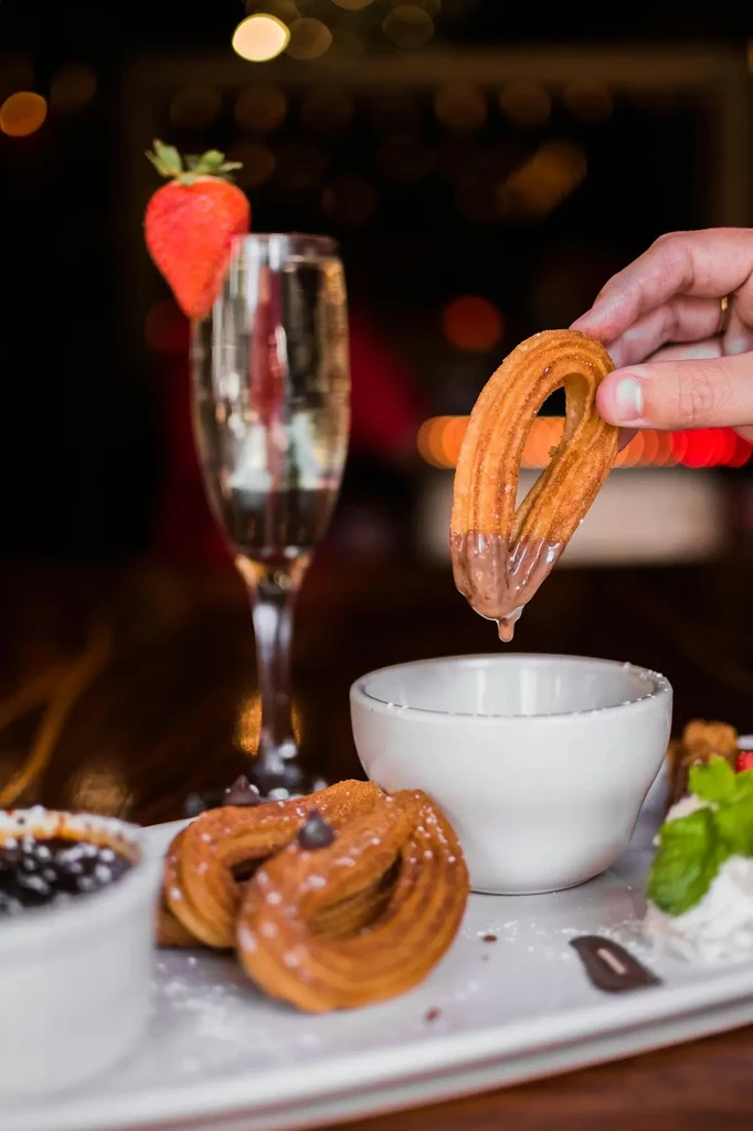 Hand dips a churro into sauce, with other desserts and a drink.