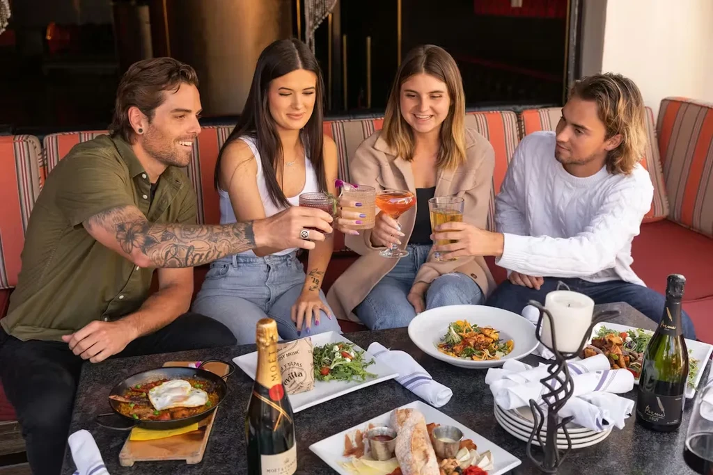 Four smiling young adults toast with drinks and food on a patio.