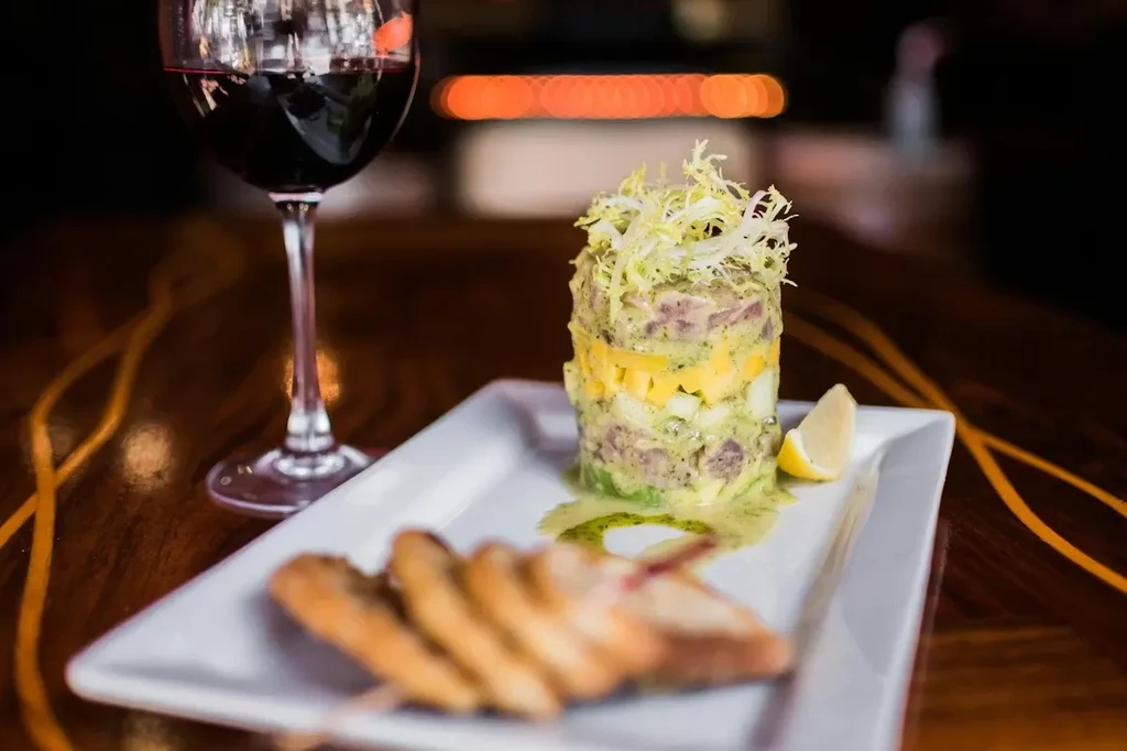 Stacked tartare, frisée, lemon, crostini, and red wine on wood.