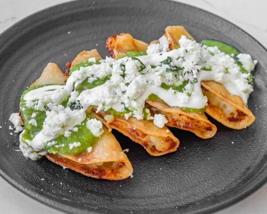 Four golden tacos/taquitos with green sauce, cream, and crumbled cheese.