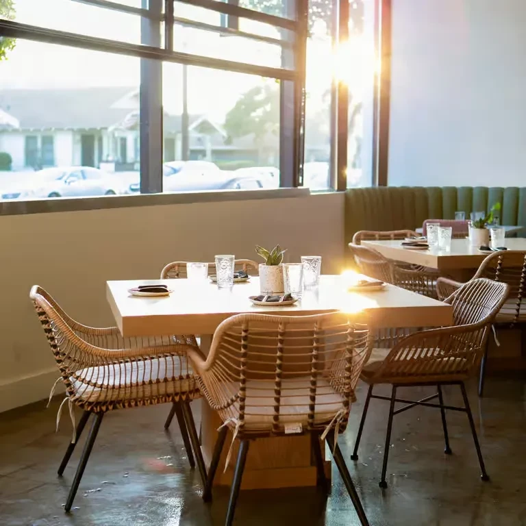 Modern restaurant dining area with sunlit tables, wicker chairs, and large windows.