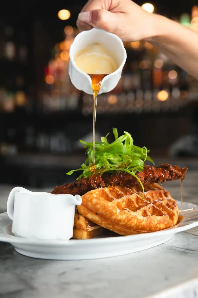 A hand pours golden syrup over crispy chicken and waffles.