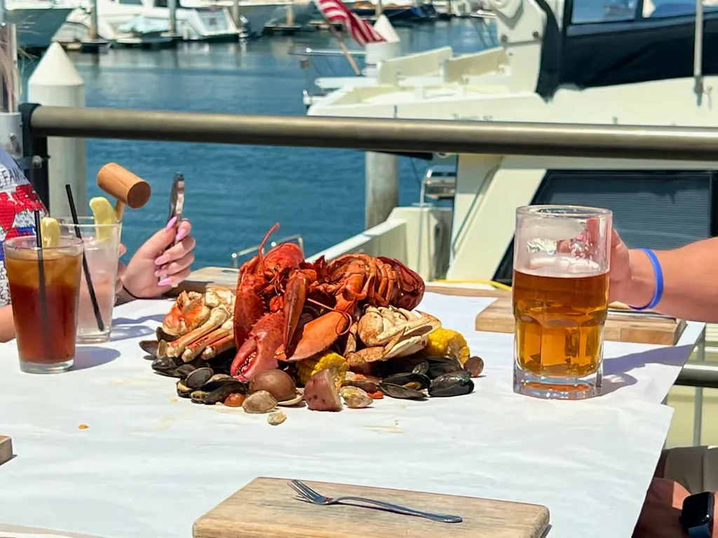 Seafood boil with lobsters, crabs, corn, and drinks at a marina.