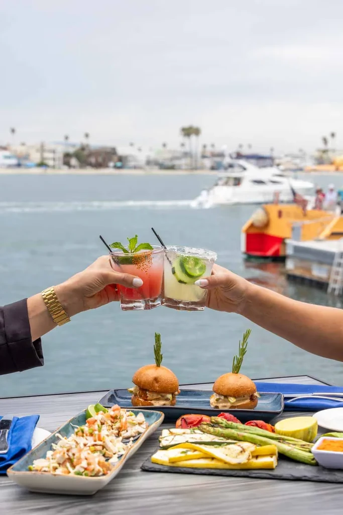 Two hands toast cocktails with appetizers and a waterfront view.
