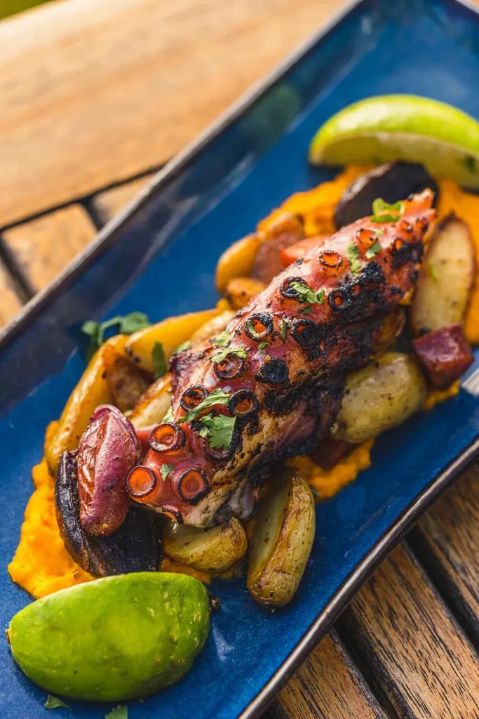 Grilled octopus tentacle with purée, potatoes, and lime on blue plate.