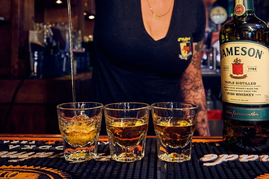 Bartender pouring whiskey into three shot glasses with Jameson bottle.