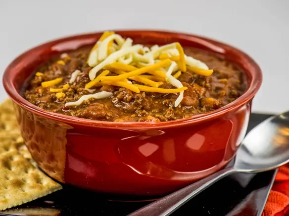 Steaming chili in red bowl, topped with cheese, crackers, and spoon.