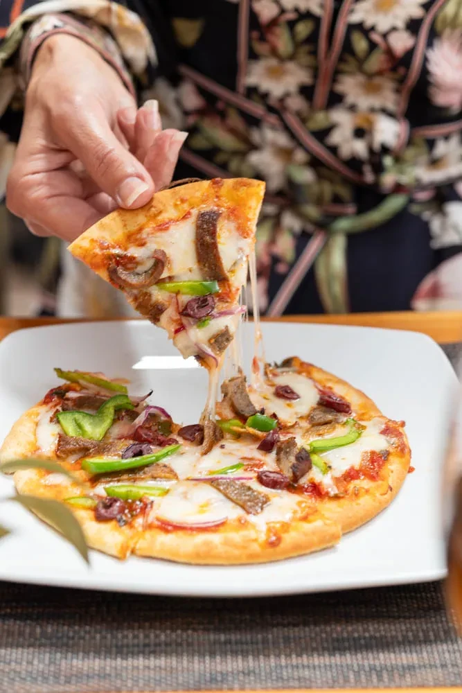 Hand lifts a slice of personal pizza with stretching cheese and various toppings.