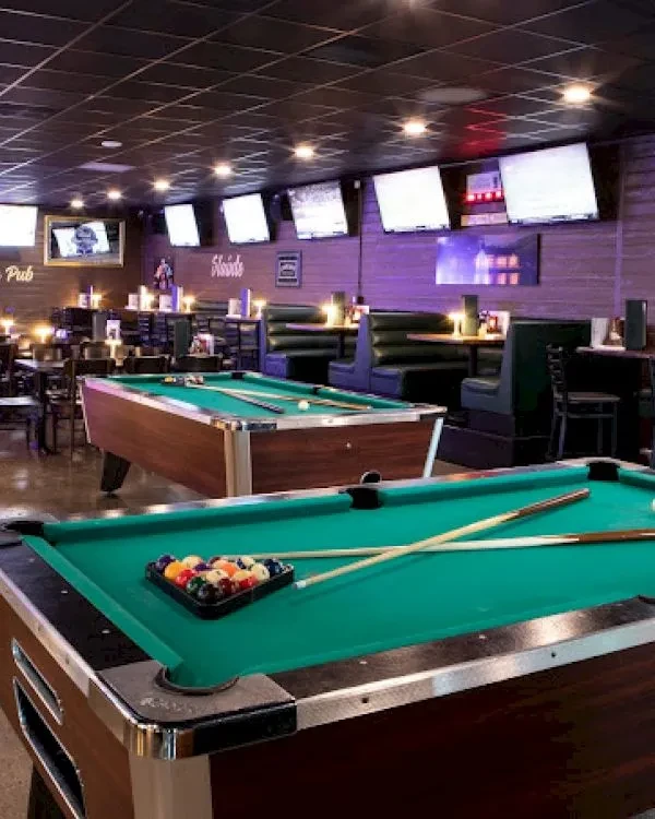 Two brightly lit pool tables in a purple-hued pub with TVs.