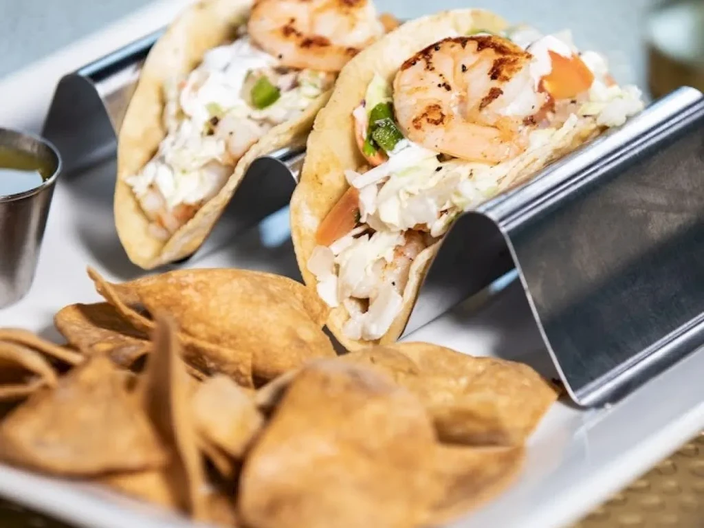 Two grilled shrimp tacos, chips, and green sauce on a plate.