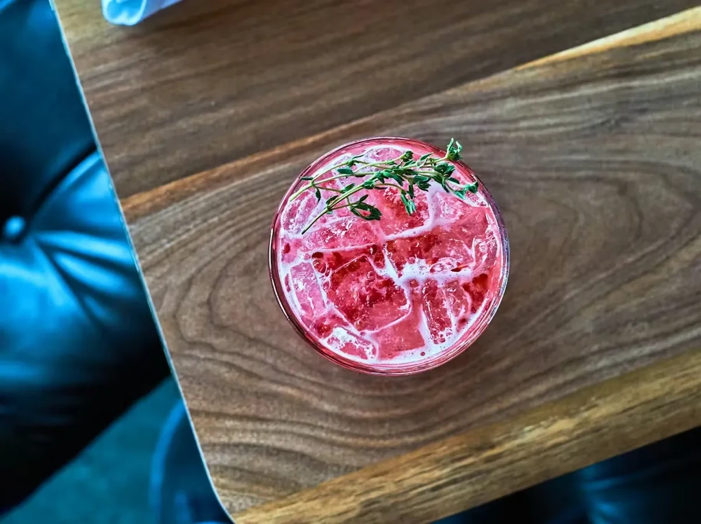 Top-down of red cocktail with ice, thyme garnish, on wood table.