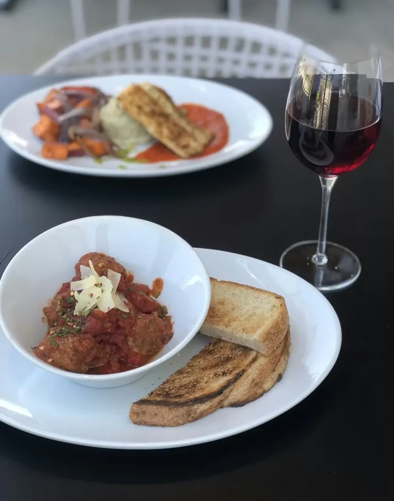 Close-up of meatballs, bread, red wine, and roasted vegetables on a dark table.