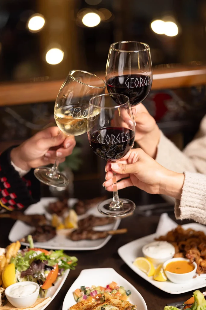 Three wine glasses etched "GEORGES" clink over a laden dinner table.