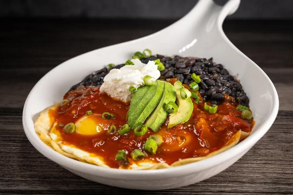 White bowl of huevos rancheros with eggs, salsa, beans, and avocado.