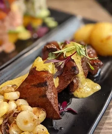 Close-up of grilled meat with creamy sauce and vegetables on a black plate.
