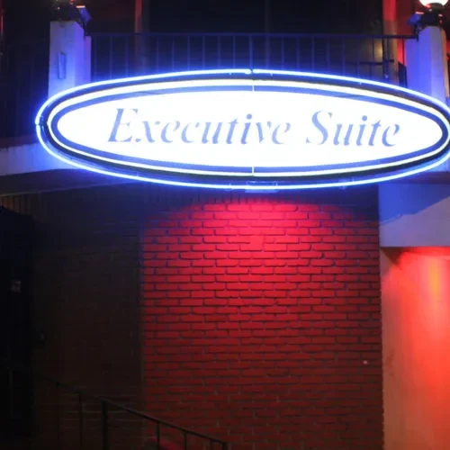 Oval neon sign "Executive Suite" illuminates a red-lit brick wall.