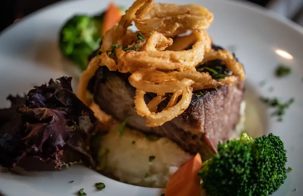 Steak topped with onion rings, mashed potatoes, and vegetables on a white plate.
