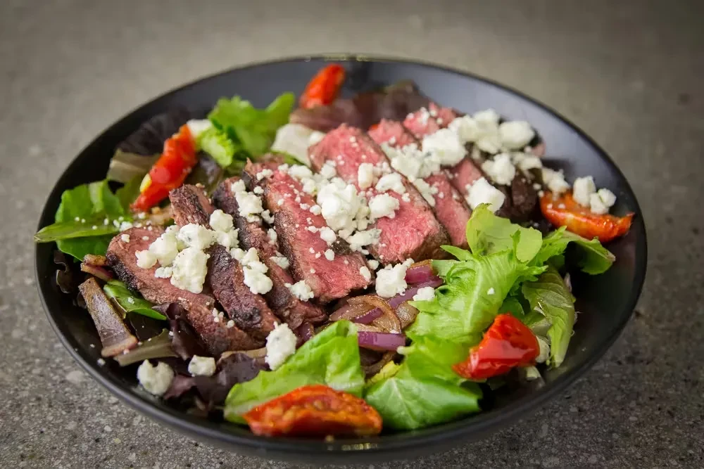 Close-up of a steak salad with medium-rare steak, cheese, and vegetables.