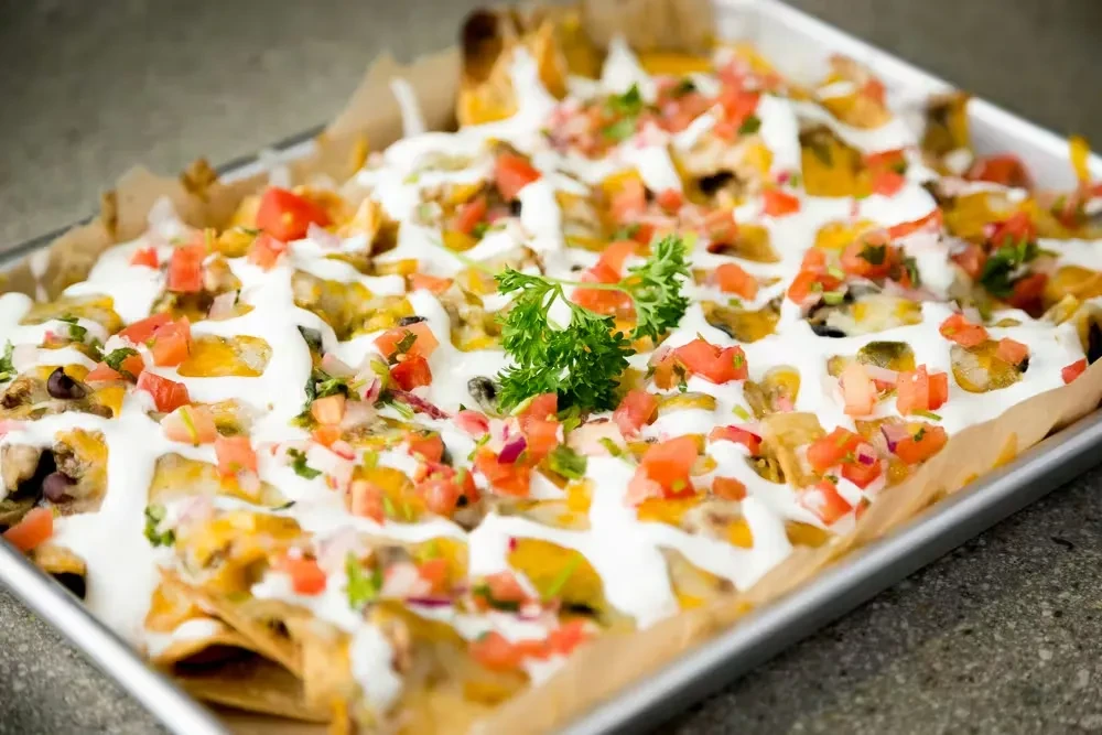 Nachos in metal tray, topped with melted cheese, vegetables, and parsley.